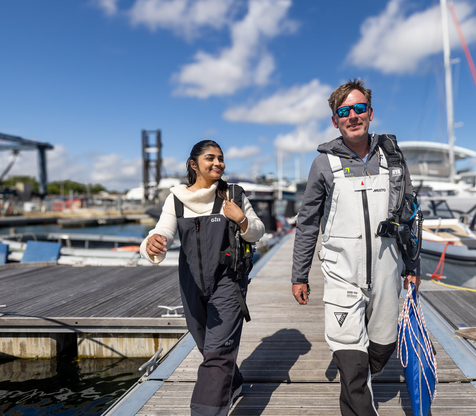 An instructor and a student in sailing kit walk down a pontoon, smiling.