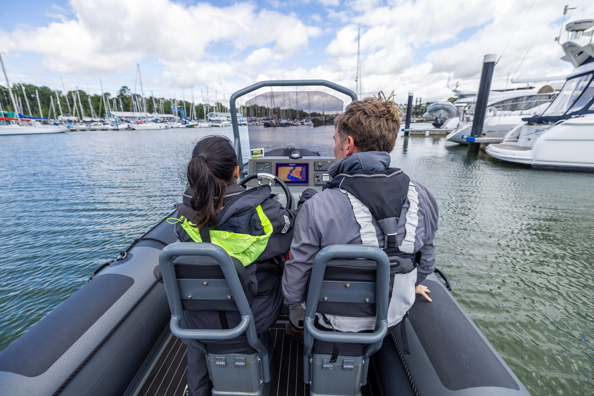 From behind: a student and her instructor at a RIB's console, heading out past moored boats.